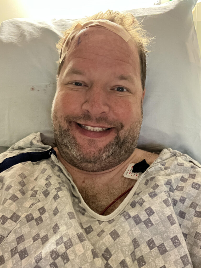 A man in a hospital gown smiles at the camera following his craniotomy to remove a benign brain tumor.