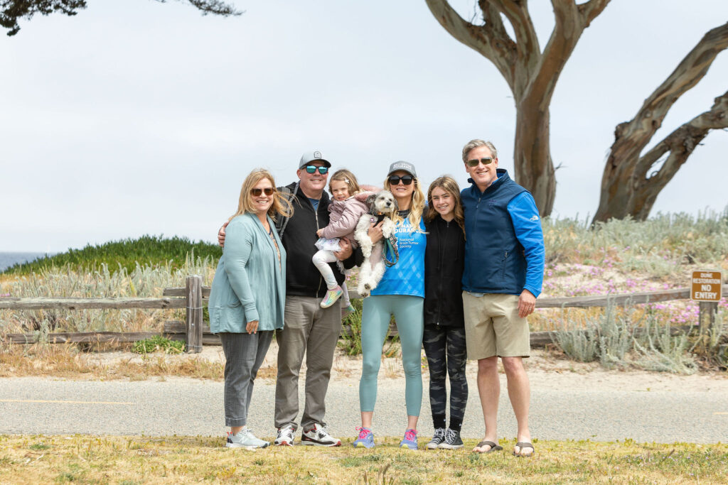 A Gray Nation Endurance runner poses for a photo with her husband, daughter, and extended family beside her. Jessica runs to raise awareness for GBM.