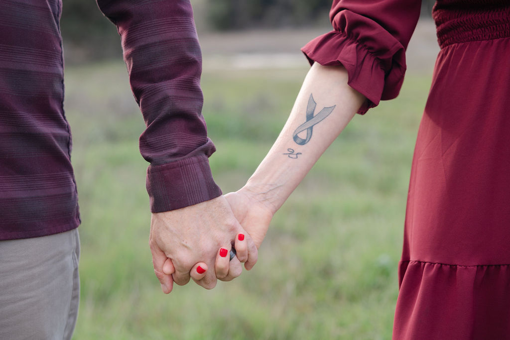 A man holds a woman's hand with a gray ribbon tattoo on her foream.