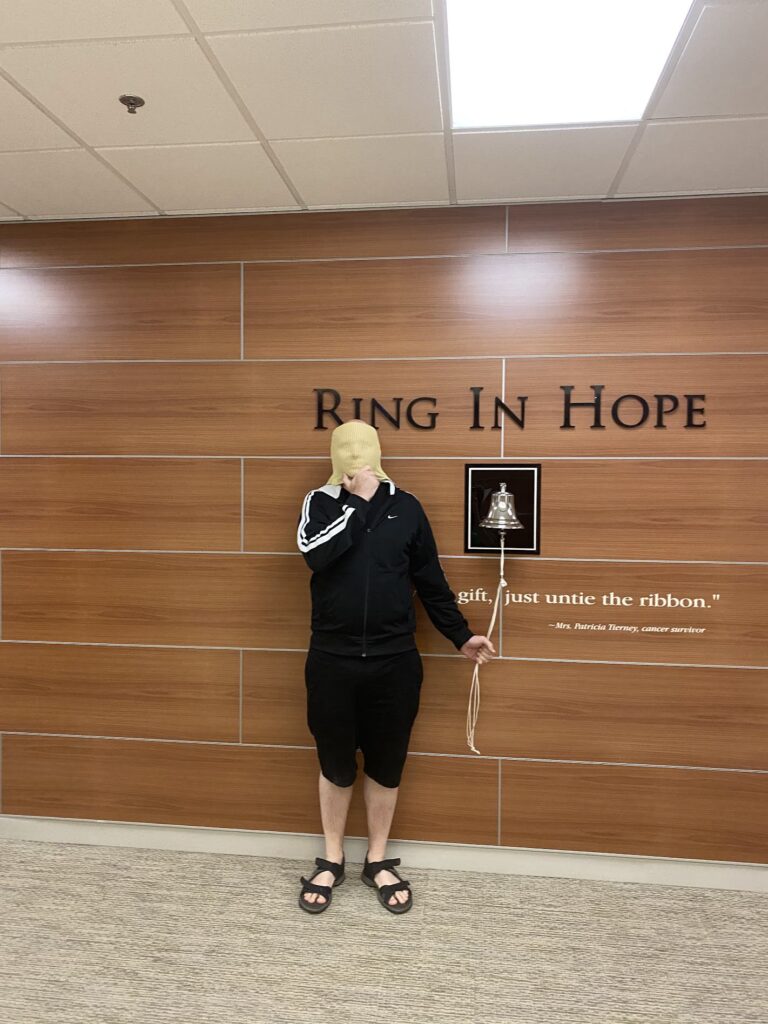 A man stands holding up a radiation mask over his head, ringing a bell to signify the end of radiation treatment.