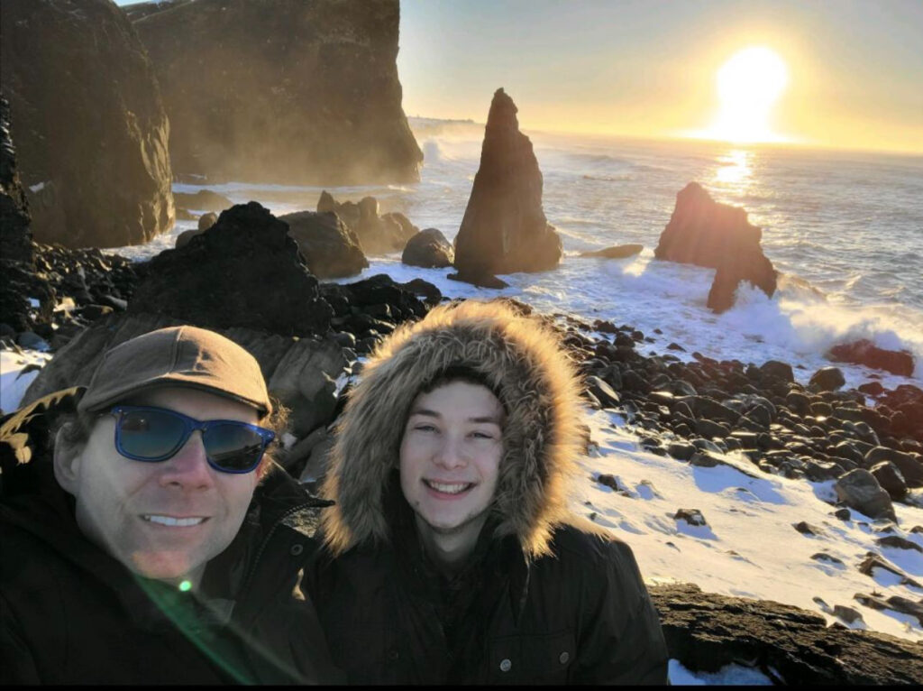 A man in a ballcap stands next to his son in a parka with a hood up. They have a blustery snowy ocean scene behind them.