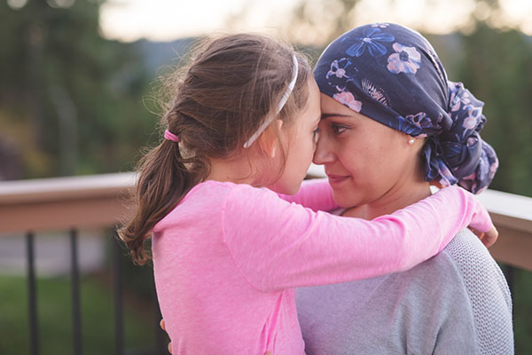A young mother parenting with a brain tumor and wearing a head scarf hugs her daughter. They have their arms around each other and they are touching noses and looking into each other's eyes. They are outside on a deck.