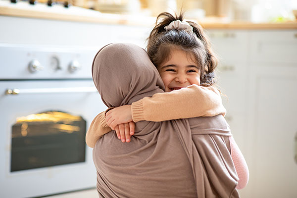 Cheerful little girl hugging tight her Muslim mom, who is parenting with a brain tumor, in hijab.