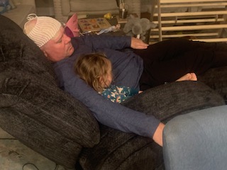 A father wearing Optune rests on a couch with his toddler daughter tucked in his arm.