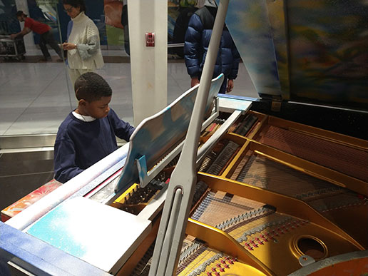 A boy in a dark blue sweatshirt with a craniotomy scar sits at a piano and plays.