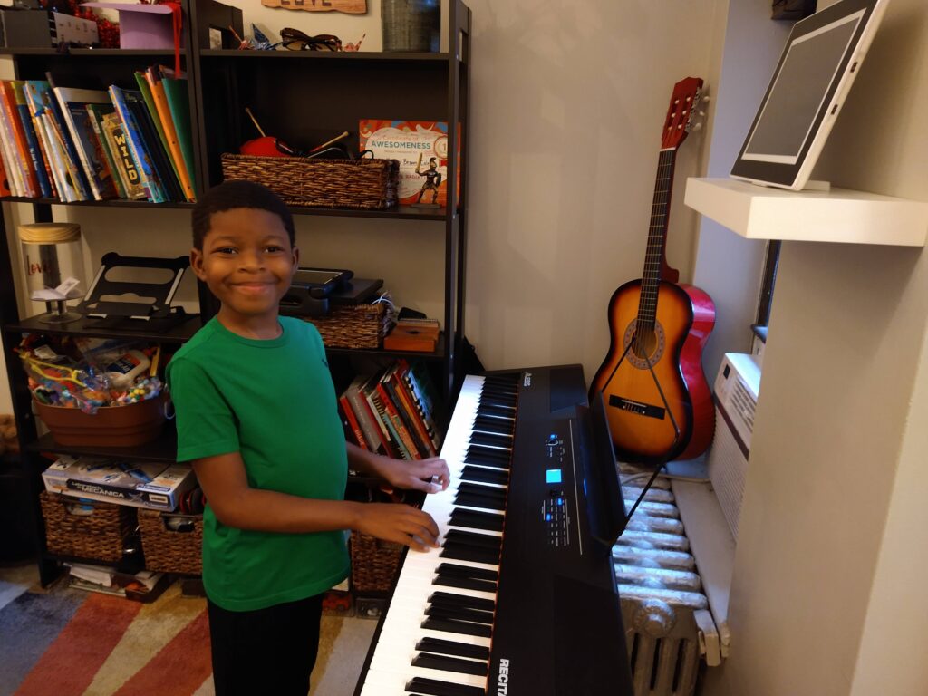 A boy in a green t-shirt stands at a keyboard with his hands resting in a position like he is about to start playing.