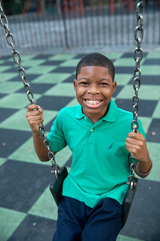 A boy, a childhood cancer patient, in a green polo sits on a swing and smiles up at the camera. 