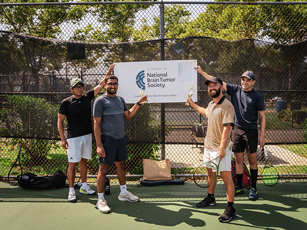 Four men in tennis gear, holding rackets, point to a banner that reads "In Support of National Brain Tumor Society" for their sports fundraiser.