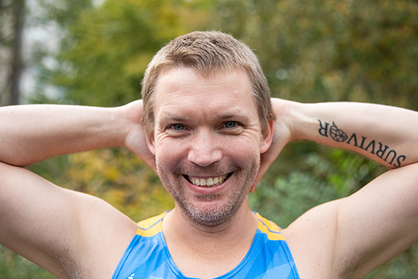 A man in a Gray Nation Endurance race bib has his hands behind his head to show off a tattoo that reads "Survivor" with a brain in place of the letter "o."