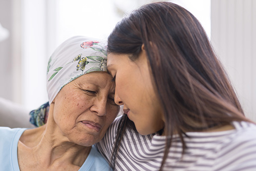An older woman with a head wrap rests her head on her daughter's shoulder. Her daughter's forehead touches her forehead.