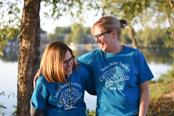 Two women in blue event t-shirts lock arms and laugh alongside a body of water.