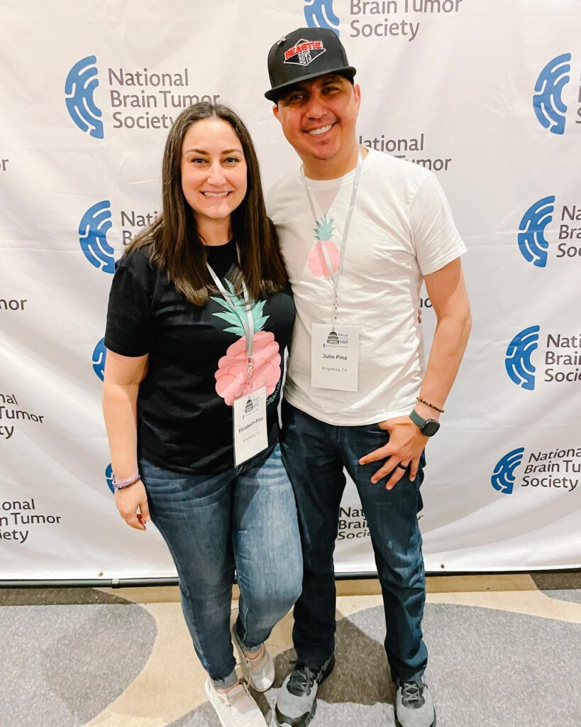 A female Caregiver Support Conversations facilitator wearing a pineapple brain shirt stands next to her husband in front of an NBTS banner.