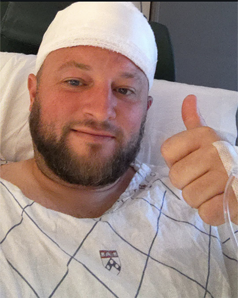 A man sits in a hospital bed giving a thumbs up with his head wrapped in a bandage following a craniotomy to remove his meningioma tumor.