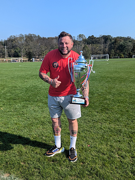 A man in a red t-shirt and gray shorts holds a trophy in one hand with a thumbs up on the other hand. He is standing on a soccer field.