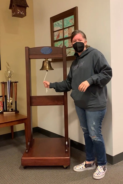 A woman, who benefited from biomarker testing, rings a bell while wearing a gray sweatshirt.