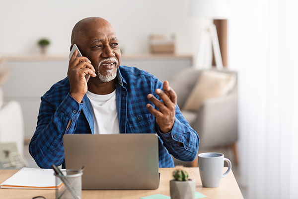 A senior man speaks on his cell phone, gesturing, from behind his laptop.