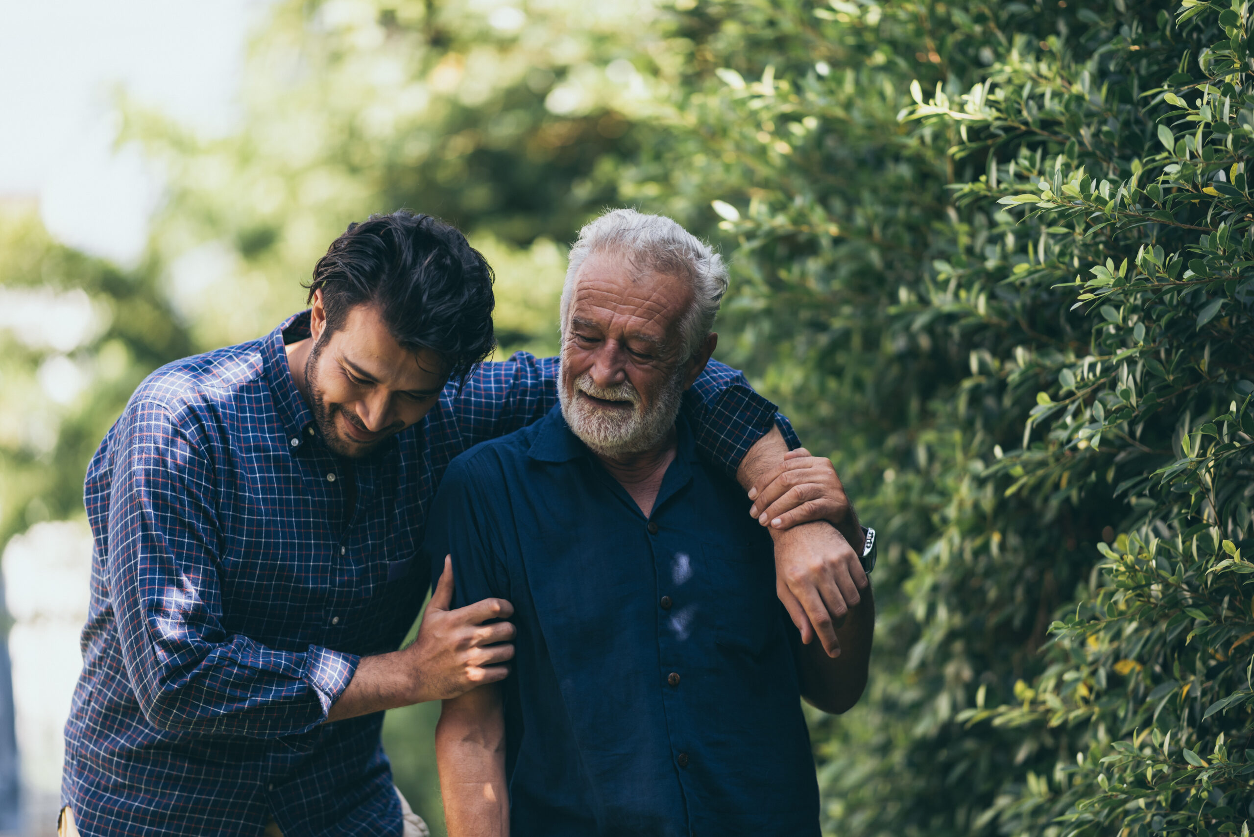 The old man and his son are walking in the park. A man hugs his elderly father, who has primary CNS lymphoma.