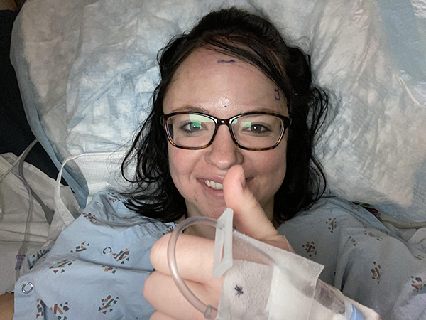 A woman in a hospital gown rests in a hospital bed while giving a thumbs up. She is in the hospital for surgery to resect a grade 2 atypical meningioma tumor.