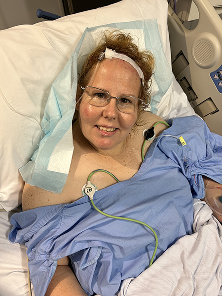 A woman rests in a hospital bed in a blue hospital gown with strips over her craniotomy scar from meningioma surgery.