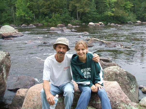 A man in a white shirt, hat, and jeans, puts his arm around his daughter in a sweatshirt and jeans.