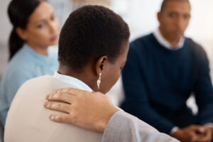An arm extends to a Black woman's shoulder as an offer of support during a support group. People with aphasia can benefit from brain tumor support groups and aphasia support groups.