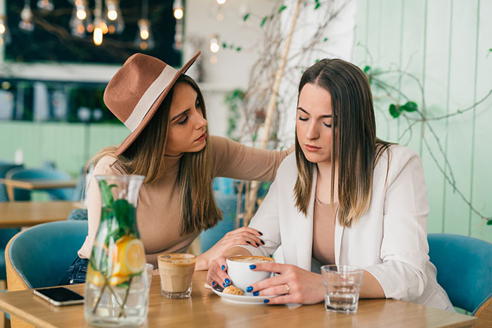 Young caucasian woman comforts her friend, who has a benign brain tumor, in cafe during coffee break.