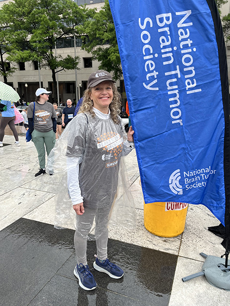 A woman in a gray t-shirt, gray hat, and clear poncho stands next to a teardrop flag that reads National Brain Tumor Society.