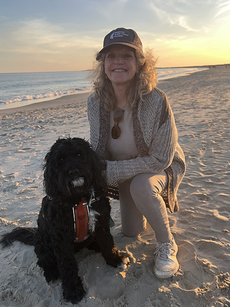 A woman kneels down by her black dog on a sandy beach with a hat that reads National Brain Tumor Society.
