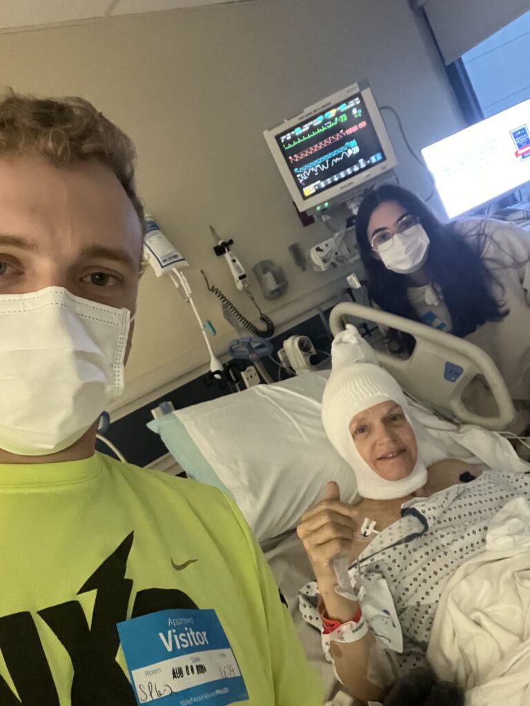 A woman with a head bandage following a craniotomy to remove meningioma smiles with her son and son's girlfriend by her hospital bed.