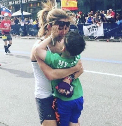 A young boy with DIPG holds his hands up to his mother's cheeks after she completed a marathon. Both wear athletic gear.