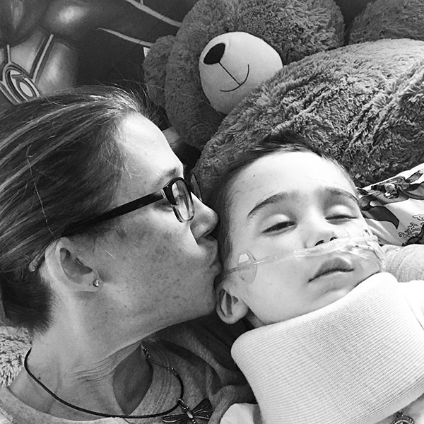 A mom in glasses kisses her boy, who has DIPG, on the side of his head while he rests with a neck brace.