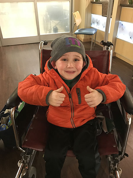 A young boy in an orange jacket and gray beanie gives a thumbs up while sitting in a wheelchair.
