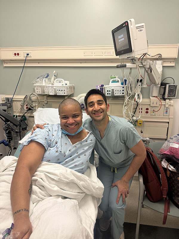 A woman with a shaved head sits in a hospital bed smiling, with her neurosurgeon in seagreen scrubs has his arm around her shoulder and smiles for the picture after a meningioma diagnosis.
