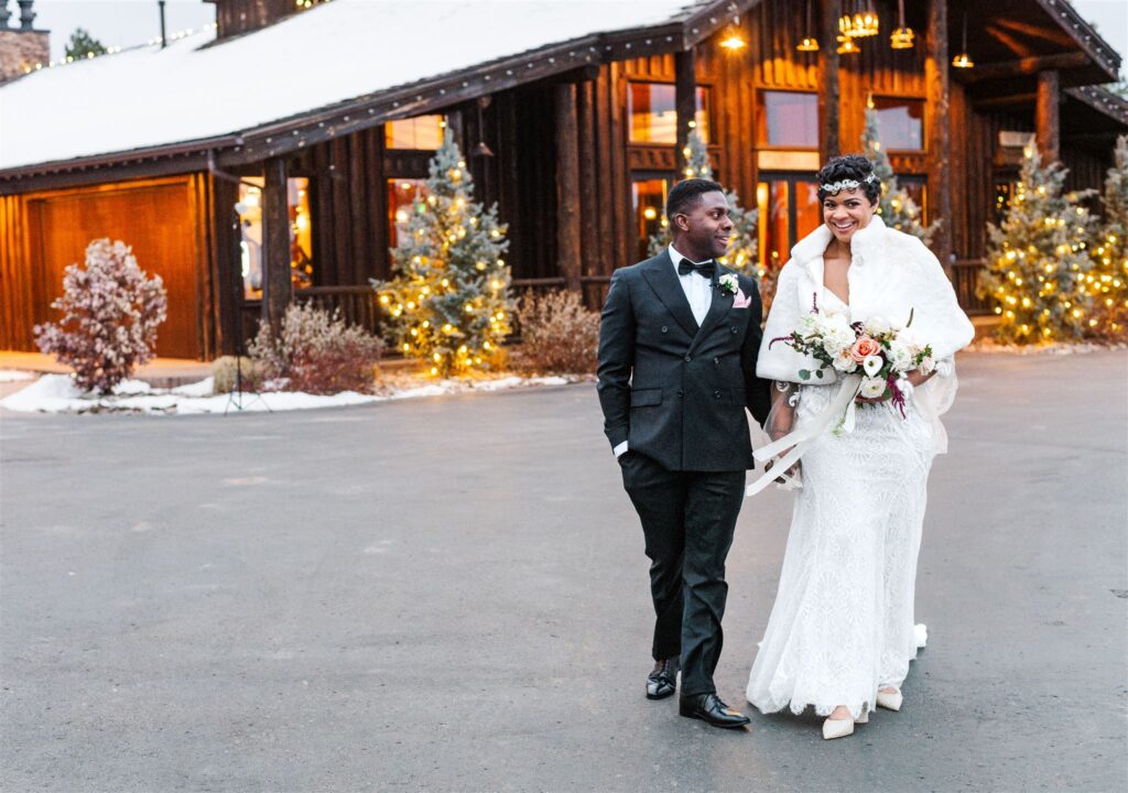 A man in a tux and a women in a white wedding dress with a fur shawl holding flowers walk with a log building behind them.