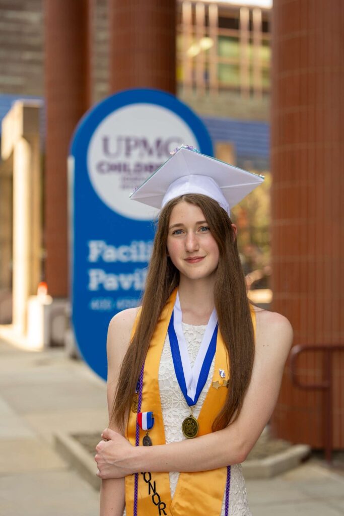 A woman in graduation garb stands outside a health center smiling.