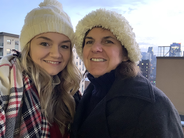 A young woman in a plaid scarf and beige winter hat stands next to her mother in a coat and hat.