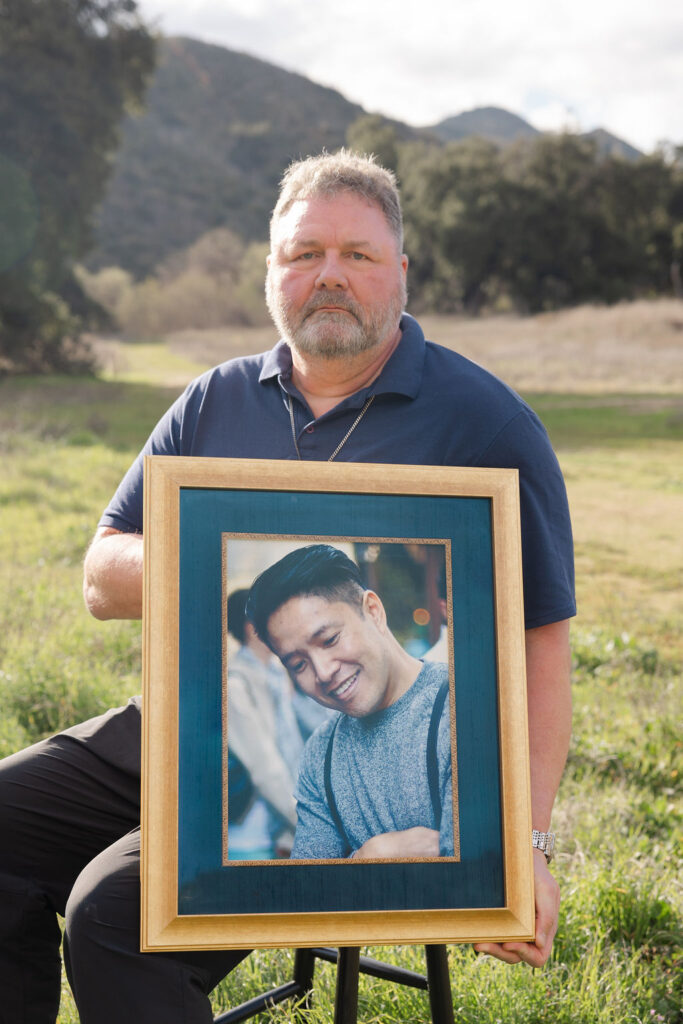 A man holds a framed photo of his husband, who passed from glioblastoma, while sitting on a stool with mountains behind him.