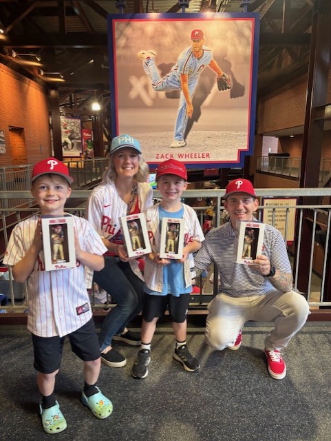 A family consisting of two young boys, a mom, and a dad wear Phillies hats while they each hold up a boxed toy.