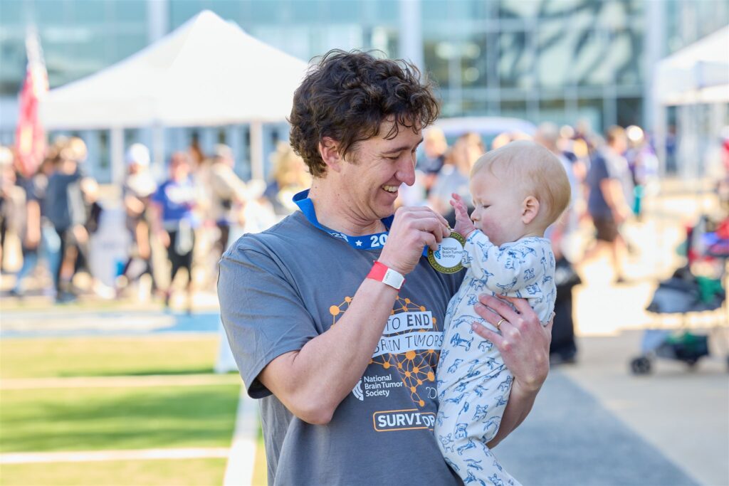 A white man shows his race medal to his 1-year-old son after the DFW Brain Tumor Walk & Race.