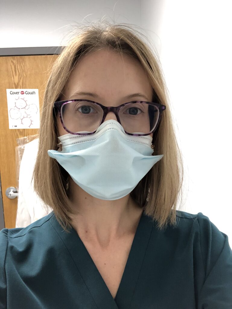 A woman wearing glasses, a mask, and scrubs looks at the camera.