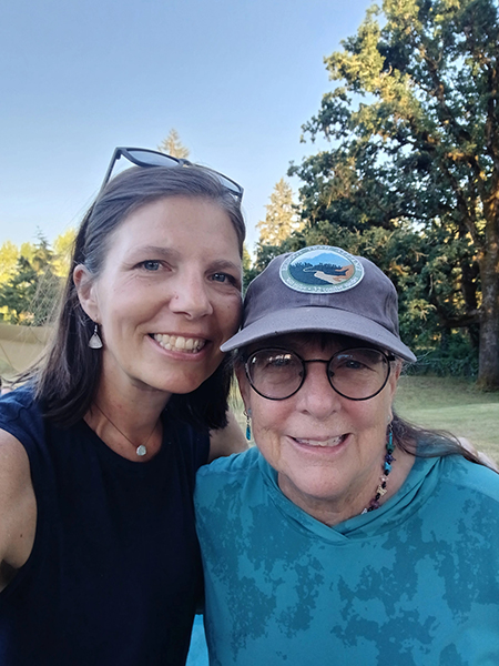 A woman, who has metastatic lung cancer, smiles alongside a friend in a teal shirt, glasses, and hat.