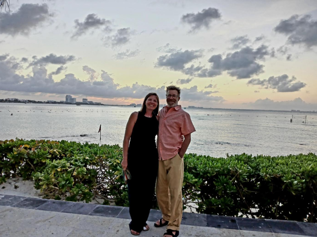 A woman, who has metastatic lung cancer, is a sleeveless black shirt and pants stands with her arm around her husband in a salmon shirt and khakis with water behind then.