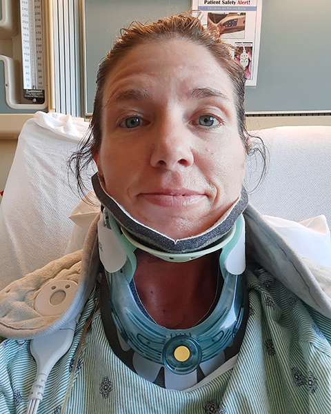 A woman, who has metastatic lung cancer, sits upright in a hospital bed wearing a C-collar and hospital gown.