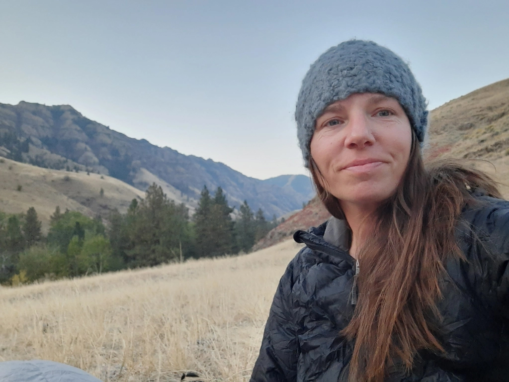 A woman, who has metastatic lung cancer, looks at the camera wearing a beanie and black jacket with a field and mountains behind her.