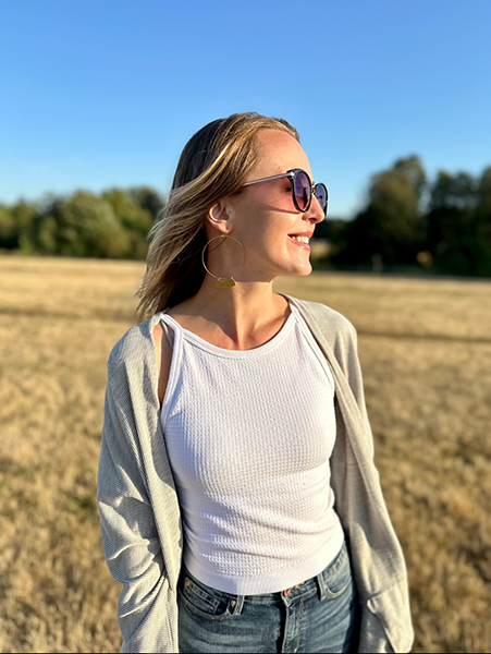 A woman with facial nerve schwannoma stands in a field on a sunny day wearing sunglasses, white shirt, jeans, and a lightweight jacket. She looks off to the side.