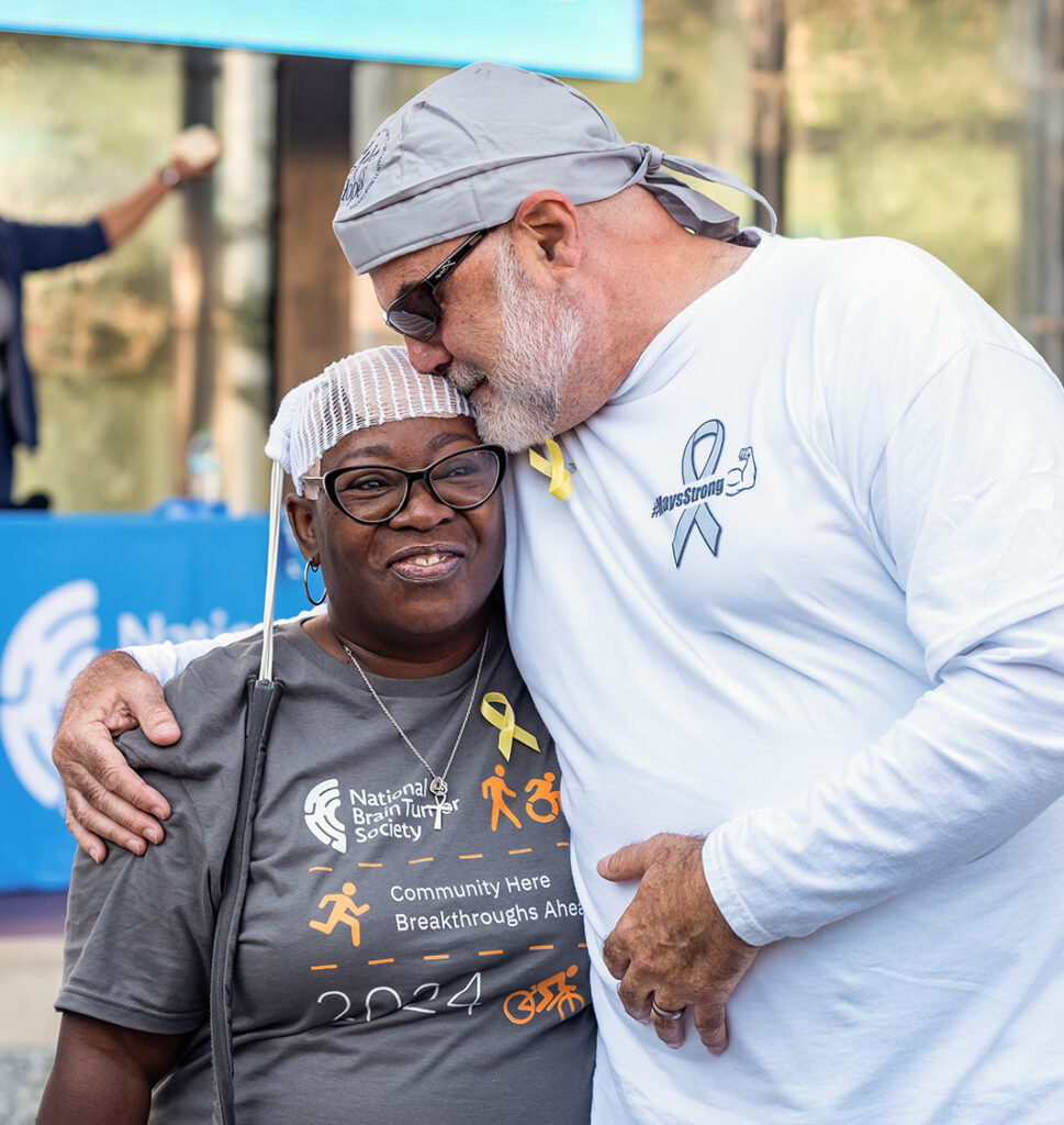 A man in a white shirt hugs a woman, who was diagnosed with glioblastoma because of nonstop babbling, wearing an NBTS t-shirt and Optune device.