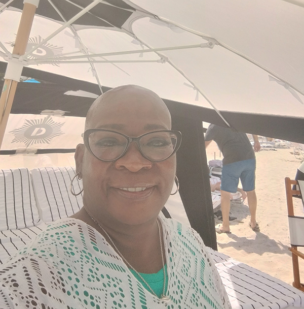 A woman, who has no hair due to glioblastoma treatment, with glasses sits under an umbrella on the beach. 