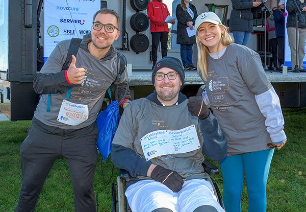 Three people in gray brain tumor survivor shirts pose for a photo at the New England Brain Tumor Walk. The middle person sits in a wheelchair. The man on the left gives a thumbs up.