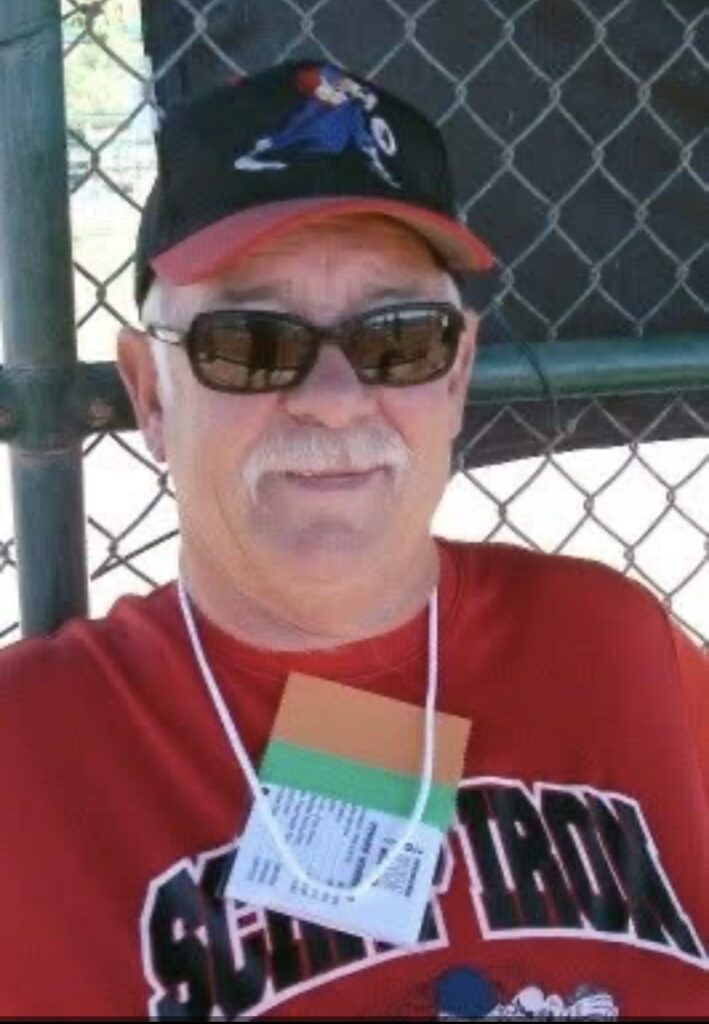 A man sits in front of a chain-linked fence wearing a hat, sunglasses, and red shirt.