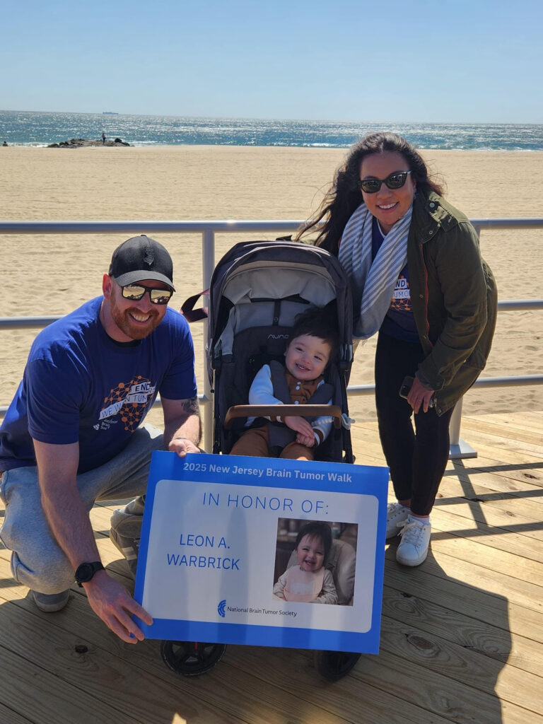 Parents wearing NBTS event t-shirts stand next to a stroller holding their 2-year-old son on a boardwalk as the father holds up a sign of his son.